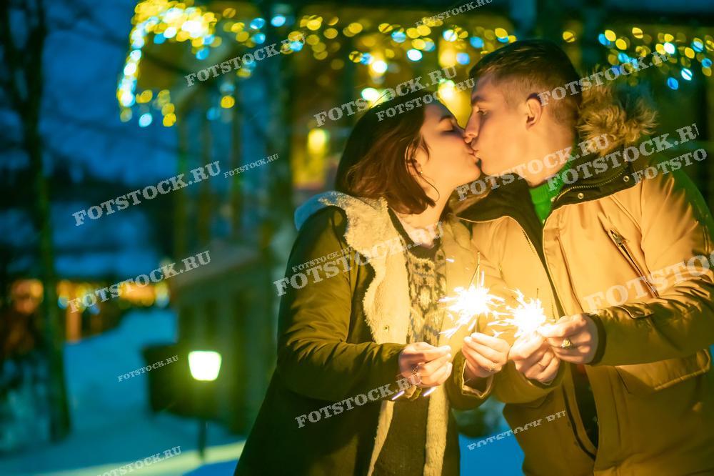 На фотографии изображена пара, целующаяся на фоне зимнего пейзажа с огоньками гирлянд в вечернее время. Оба держат в руках зажжённые бенгальские огни, создавая атмосферу праздника и уюта. Они одеты в теплую зимнюю одежду, а вокруг царит вечерняя новогодняя атмосфера с огнями и гирляндами на заднем плане.